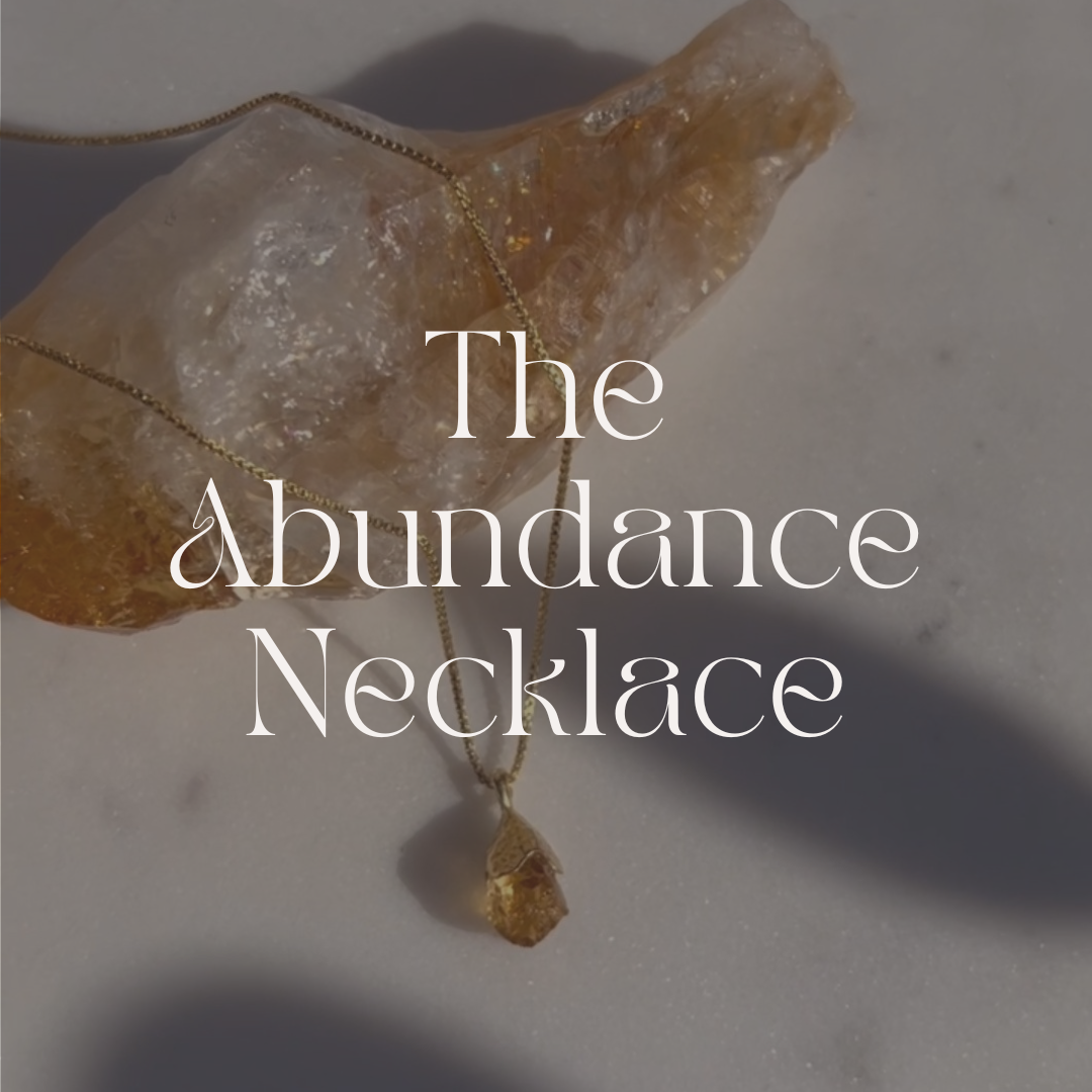 Necklace with a crystal pendant on a gray background, labeled 'The Abundance Necklace'.