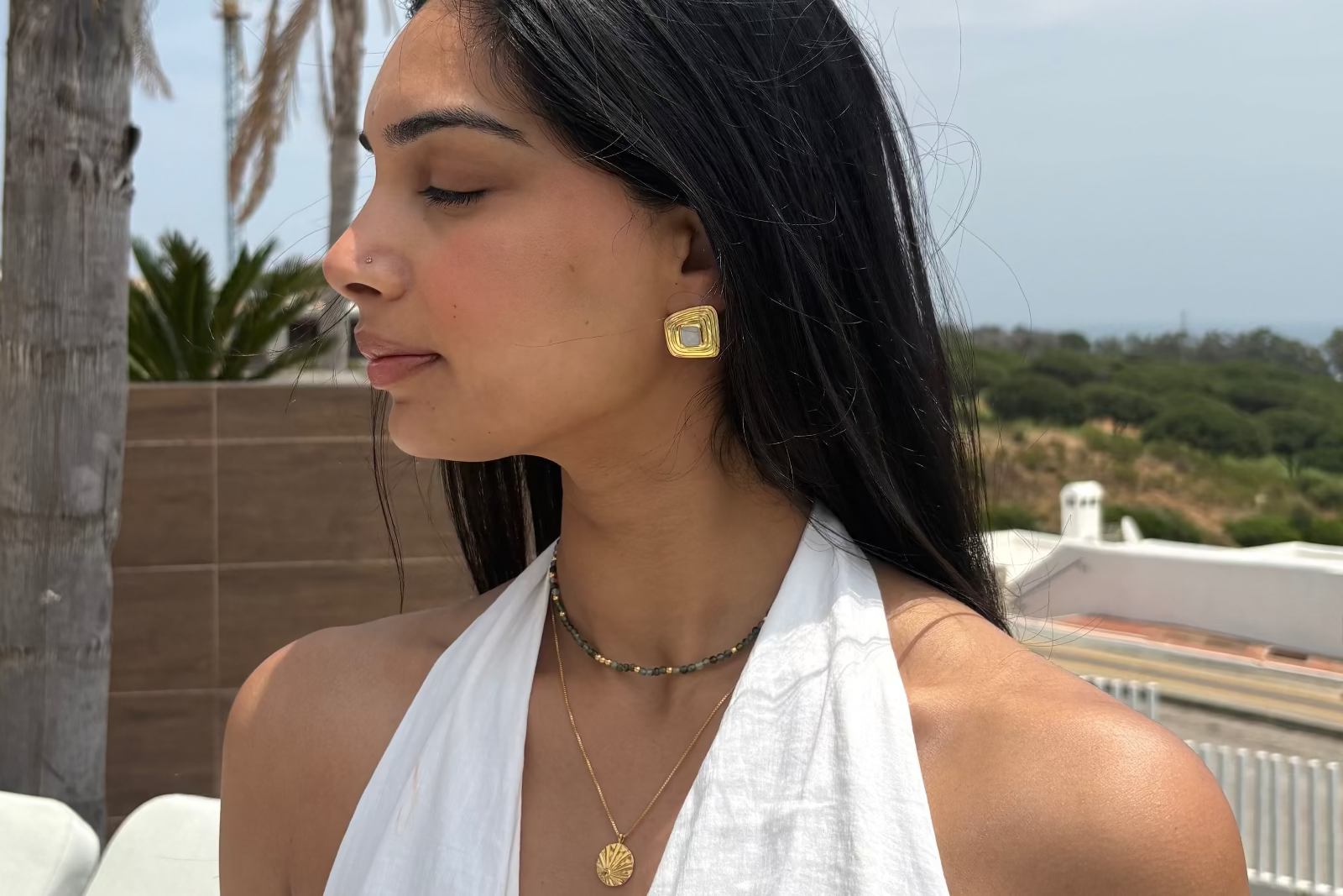 Woman wearing gold earrings and a necklace outdoors with palm trees and buildings in the background
